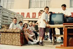 Uma fotografia colorida que mostra um grupo de crianças e jovens posando em um cenário montado ao ar livre, possivelmente em um evento comunitário ou promocional. O ambiente inclui um sofá estampado, uma mesa com uma televisão antiga e um fundo com letreiros comerciais, refletindo a moda e o estilo de vida da década de 1990.