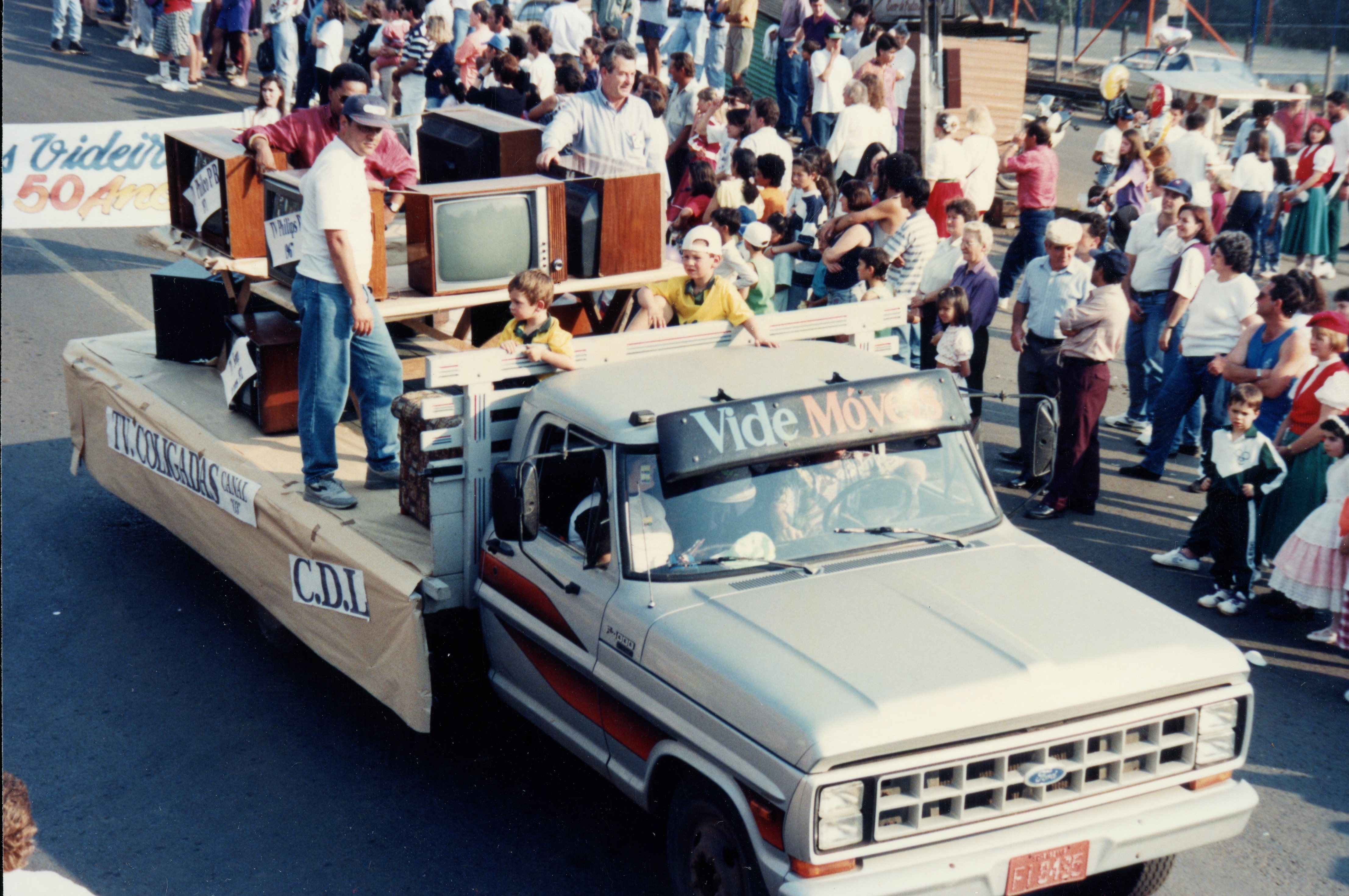 Uma fotografia de um desfile comemorativo em Videira, possivelmente celebrando o aniversário da cidade, com um caminhão Ford decorado como carro alegórico transportando televisores antigos. O veículo exibe faixas com os nomes 'Vide Móveis' e 'TV Coligadas', refletindo o contexto comercial e cultural da época, provavelmente na década de 1980 ou início de 1990.