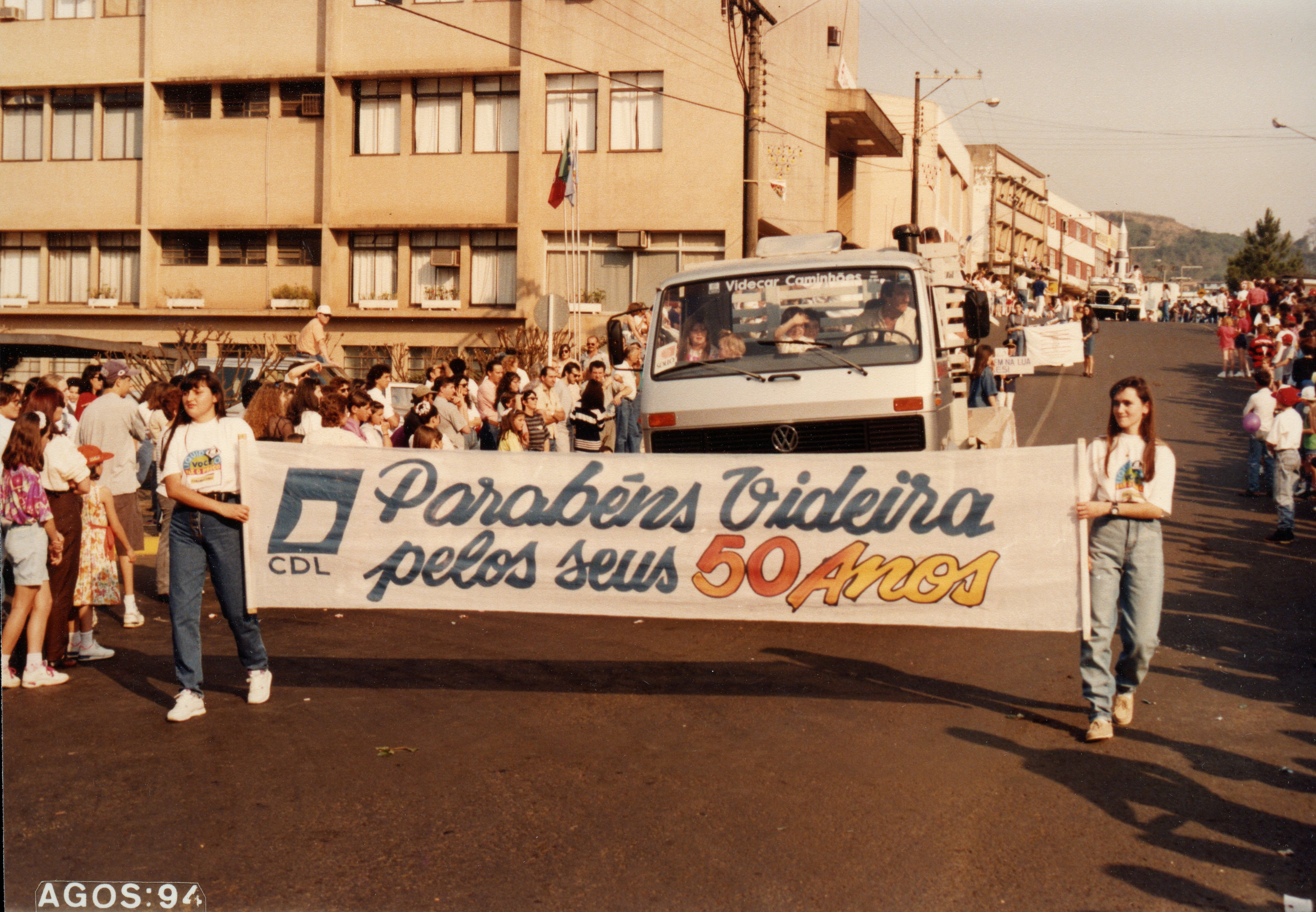 Uma fotografia colorida que registra um desfile comemorativo em Videira, Santa Catarina, em agosto de 1994. Duas jovens carregam uma faixa celebrando os 50 anos da cidade, enquanto um caminhão da Volkswagen e uma multidão acompanham o evento em uma rua urbana.