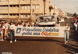 Uma fotografia colorida que registra um desfile comemorativo em Videira, Santa Catarina, em agosto de 1994. Duas jovens carregam uma faixa celebrando os 50 anos da cidade, enquanto um caminhão da Volkswagen e uma multidão acompanham o evento em uma rua urbana.