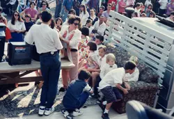 Uma fotografia colorida que retrata um evento comunitário ou celebração ao ar livre, possivelmente na década de 1990. Vê-se um grupo de pessoas, incluindo crianças sentadas em um sofá colocado sobre um tapete, com adultos ao redor e uma televisão antiga sobre uma mesa, sugerindo uma atividade recreativa ou educativa em uma praça ou espaço público.