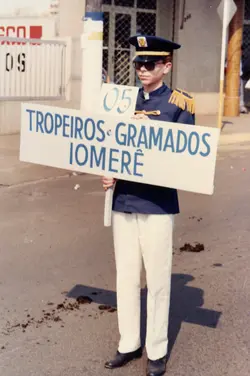 Uma fotografia de um jovem em um desfile cívico, vestindo um uniforme de banda marcial com dragonas e boné. Ele segura uma placa indicando 'Tropeiros e Gramados Iomerê', sugerindo um evento cultural ou comemorativo em uma cidade do interior catarinense, provavelmente nas décadas de 1980 ou 1990.