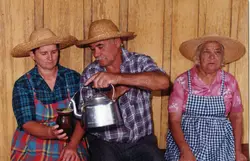 Uma fotografia colorida que retrata três pessoas vestidas com trajes típicos rurais, incluindo chapéus de palha e camisas xadrez, em um ambiente com paredes de madeira. O homem ao centro serve chimarrão, uma prática cultural comum no sul do Brasil, sugerindo uma cena de convívio familiar ou tradição regional.