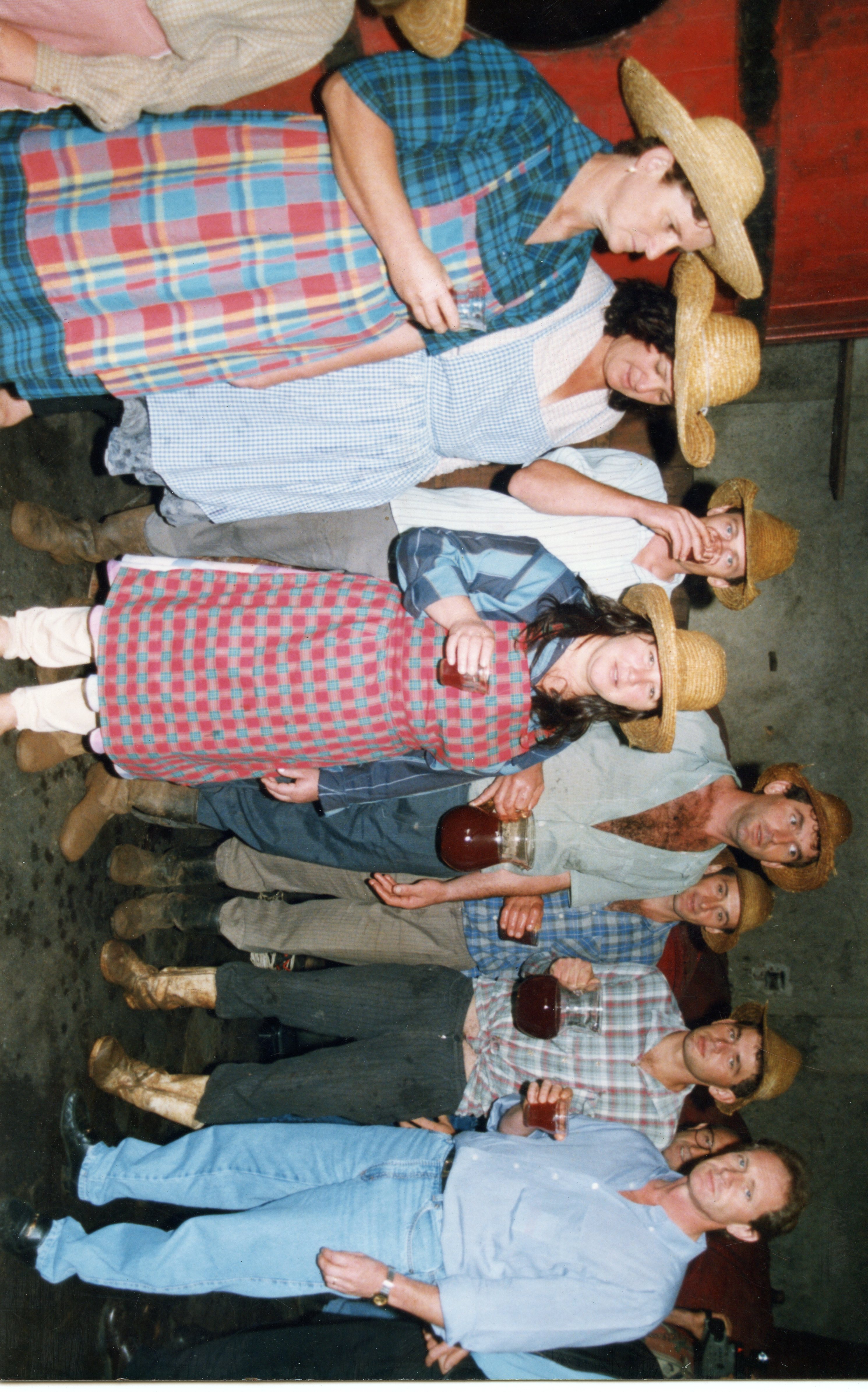 Uma fotografia de um grupo de pessoas em um ambiente rural ou de produção de vinho, possivelmente em uma celebração ou colheita. Os participantes vestem roupas simples, chapéus de palha e seguram jarras de vidro com líquido escuro, sugerindo uma atividade tradicional de vinificação ou festa típica.