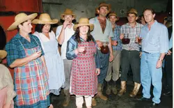 Uma fotografia de um grupo de pessoas em um ambiente rural ou de produção de vinho, possivelmente em uma celebração ou colheita. Os participantes vestem roupas simples, chapéus de palha e seguram jarras de vidro com líquido escuro, sugerindo uma atividade tradicional de vinificação ou festa típica.