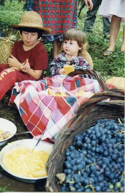Uma cena de piquenique ao ar livre, provavelmente durante uma colheita de uvas, com duas crianças sentadas no chão comendo. O ambiente sugere uma atividade rural ou familiar, possivelmente em uma região de cultivo de uvas como Videira, com cestas de vime e alimentos simples.