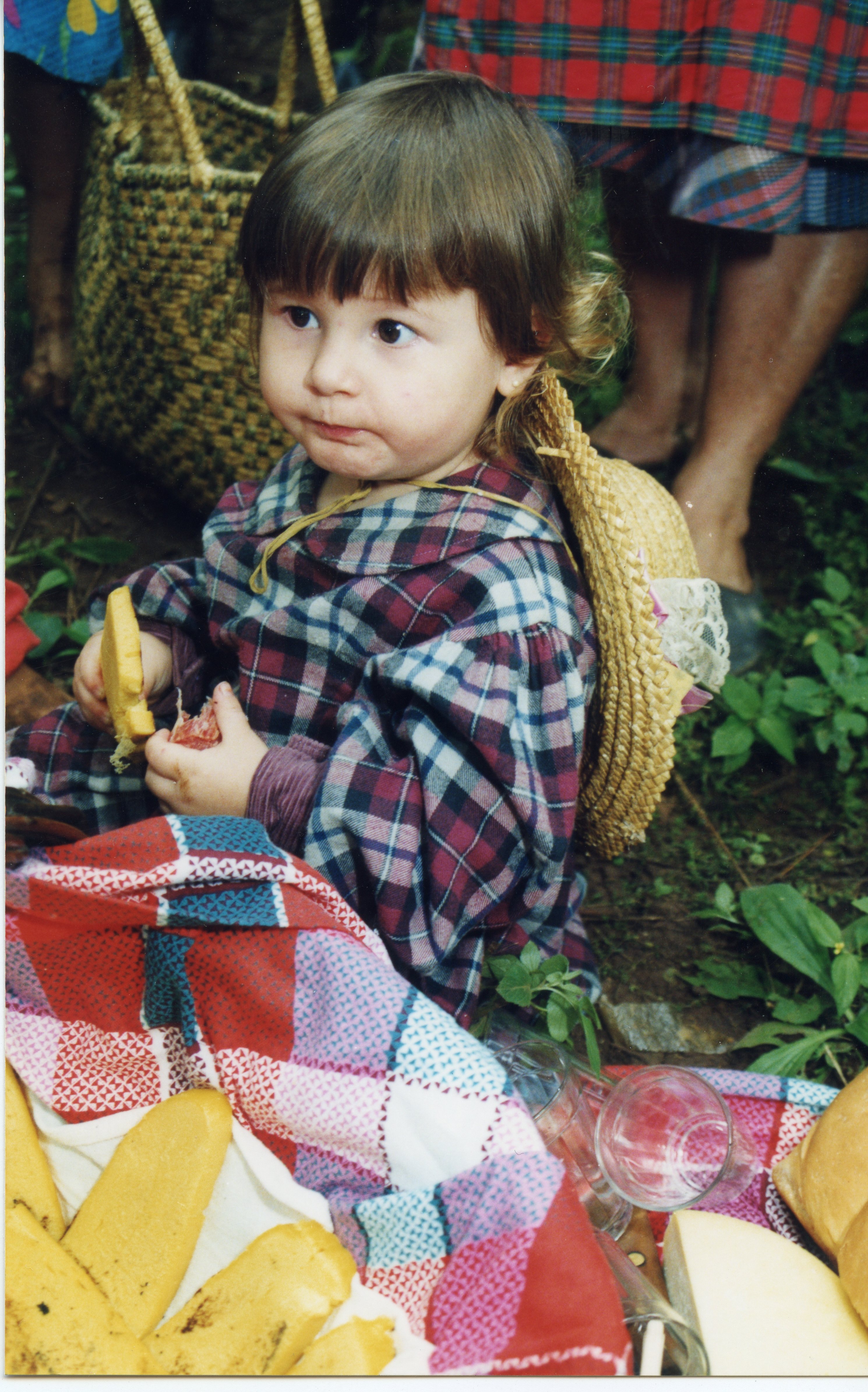 Uma criança pequena vestida com roupas de estilo caipira ou tradicional, incluindo um chapéu de palha, sentada em um piquenique ao ar livre. A cena sugere uma celebração cultural ou festividade rural, possivelmente uma festa junina, com alimentos como pães e queijos dispostos sobre uma toalha xadrez.