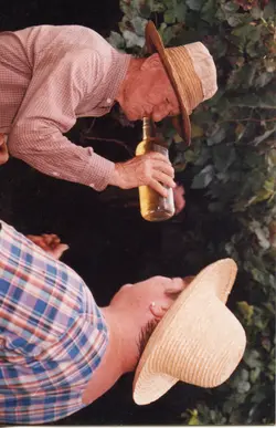 Uma fotografia colorida que retrata dois homens usando chapéus de palha, possivelmente durante uma pausa no trabalho rural ou colheita. Um dos homens bebe de uma garrafa de vidro, sugerindo um momento de descanso em um ambiente agrícola, típico da cultura de pequenas propriedades rurais brasileiras.