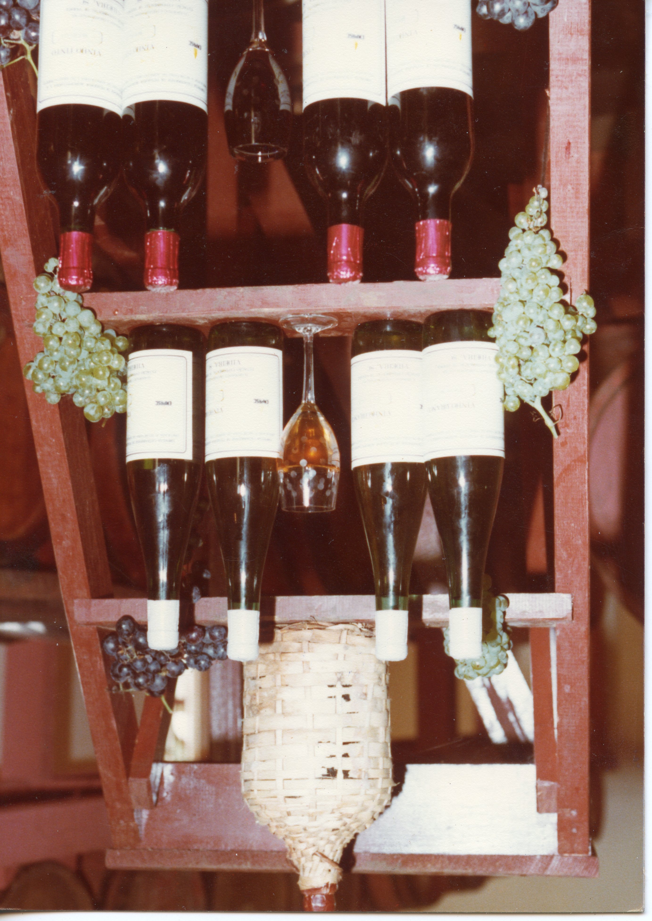A imagem exibe uma prateleira de madeira rústica decorada com garrafas de vinho, taças e cachos de uvas, sugerindo um ambiente de vinícola ou adega. A composição evoca a tradição vitivinícola, possivelmente associada à cultura de cidades como Videira, destacando a produção local de vinhos.
