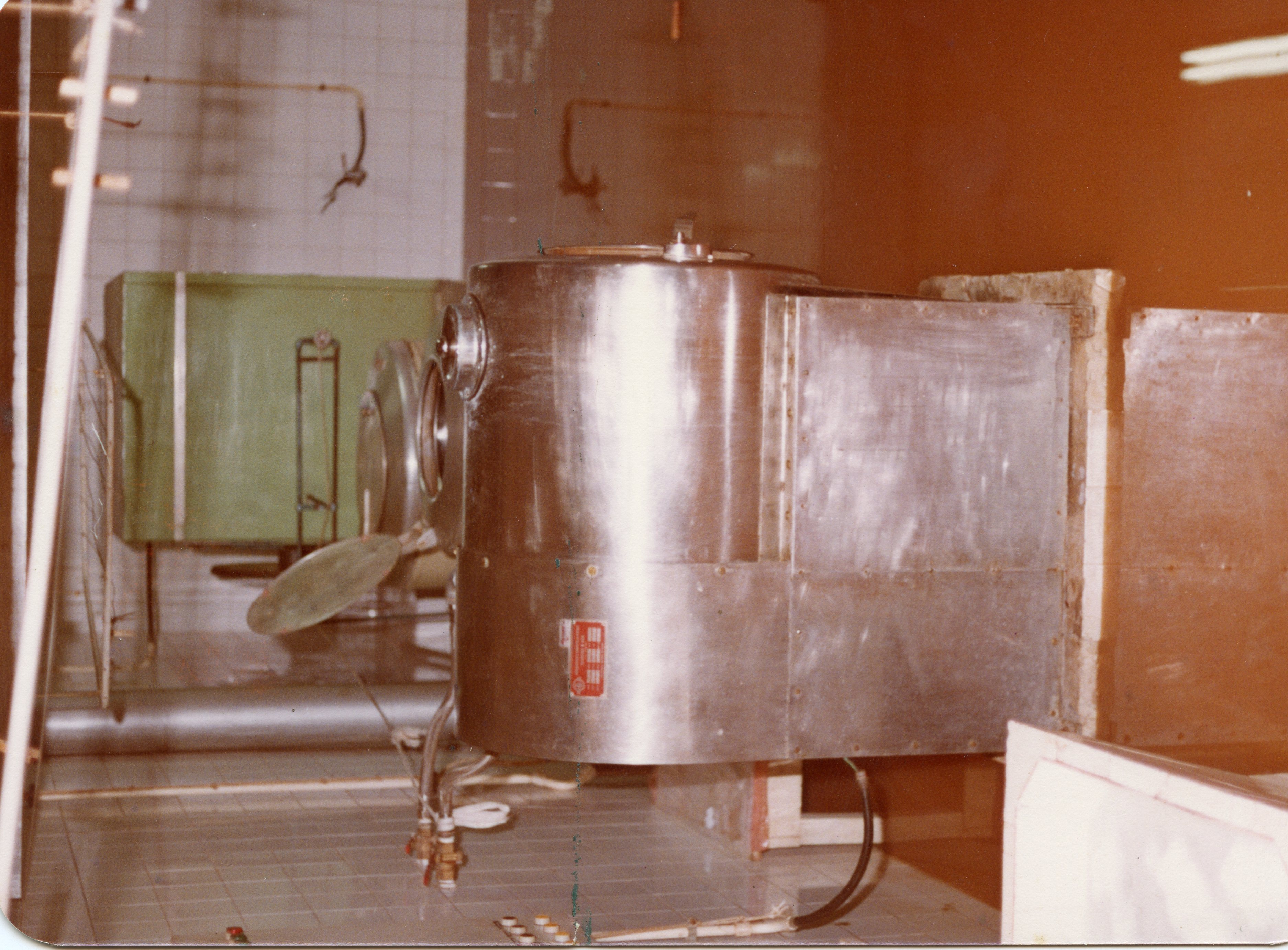 A imagem mostra o interior de uma instalação industrial ou laboratorial, possivelmente uma fábrica de alimentos ou laticínios, com um grande tanque de aço inoxidável em destaque. O ambiente apresenta azulejos brancos e equipamentos de processamento, sugerindo um contexto de produção industrial da segunda metade do século XX.