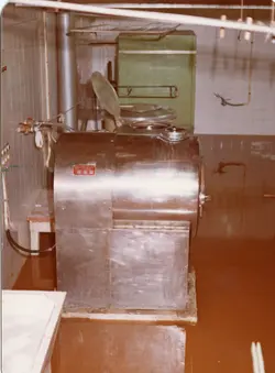 A imagem mostra o interior de uma instalação industrial ou laboratorial, possivelmente uma fábrica de alimentos ou laticínios, com um grande tanque de aço inoxidável em destaque. O ambiente apresenta azulejos brancos e equipamentos de processamento, sugerindo um contexto de produção industrial da segunda metade do século XX.