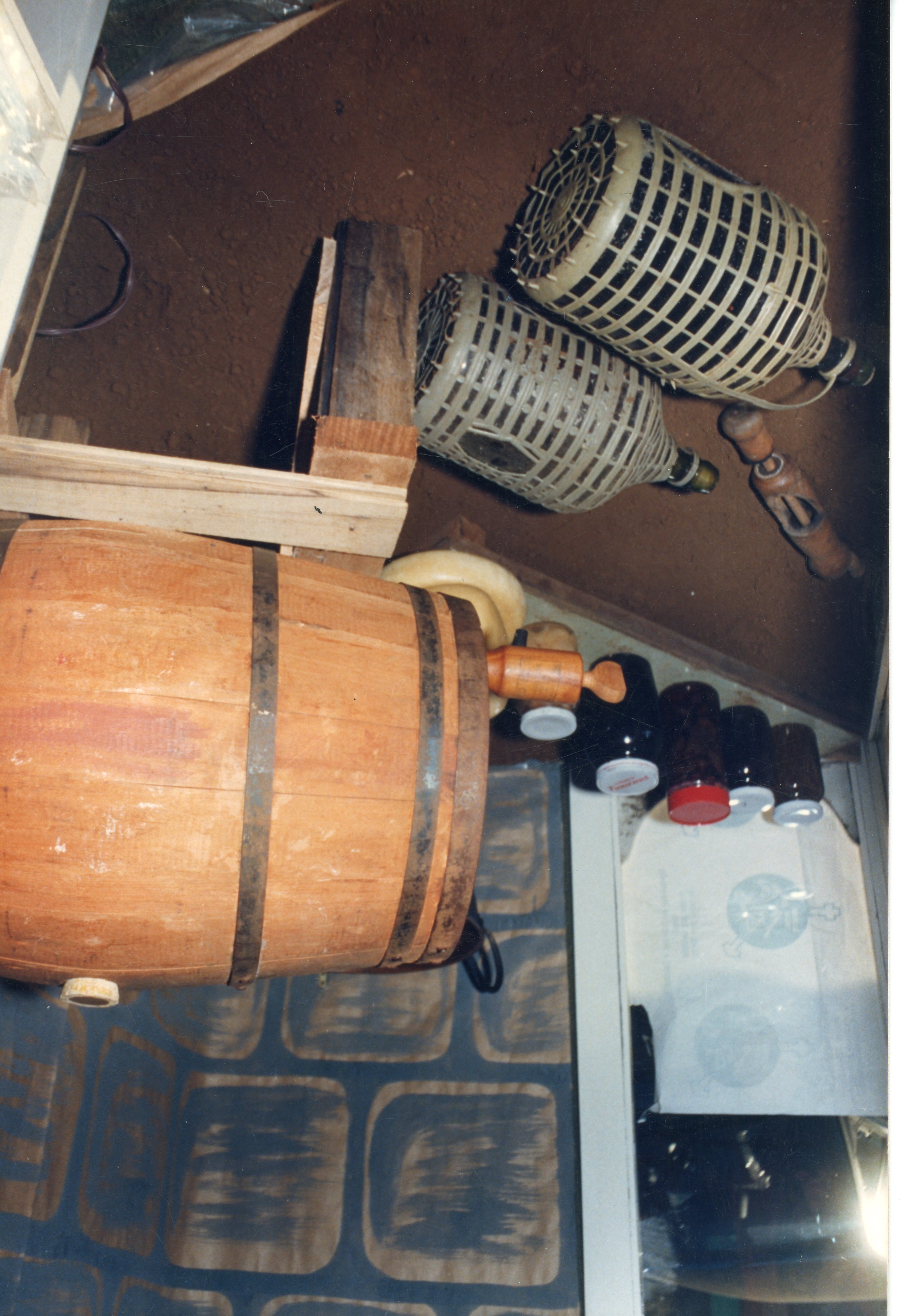 A imagem mostra um arranjo de objetos tradicionais de produção artesanal, incluindo um barril de madeira, garrafões revestidos de palha e potes de vidro. O cenário sugere um contexto de produção vinícola ou de conservas caseiras, comum em regiões de colonização europeia no Brasil, como Videira.