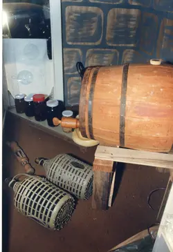 A imagem mostra um arranjo de objetos tradicionais de produção artesanal, incluindo um barril de madeira, garrafões revestidos de palha e potes de vidro. O cenário sugere um contexto de produção vinícola ou de conservas caseiras, comum em regiões de colonização europeia no Brasil, como Videira.