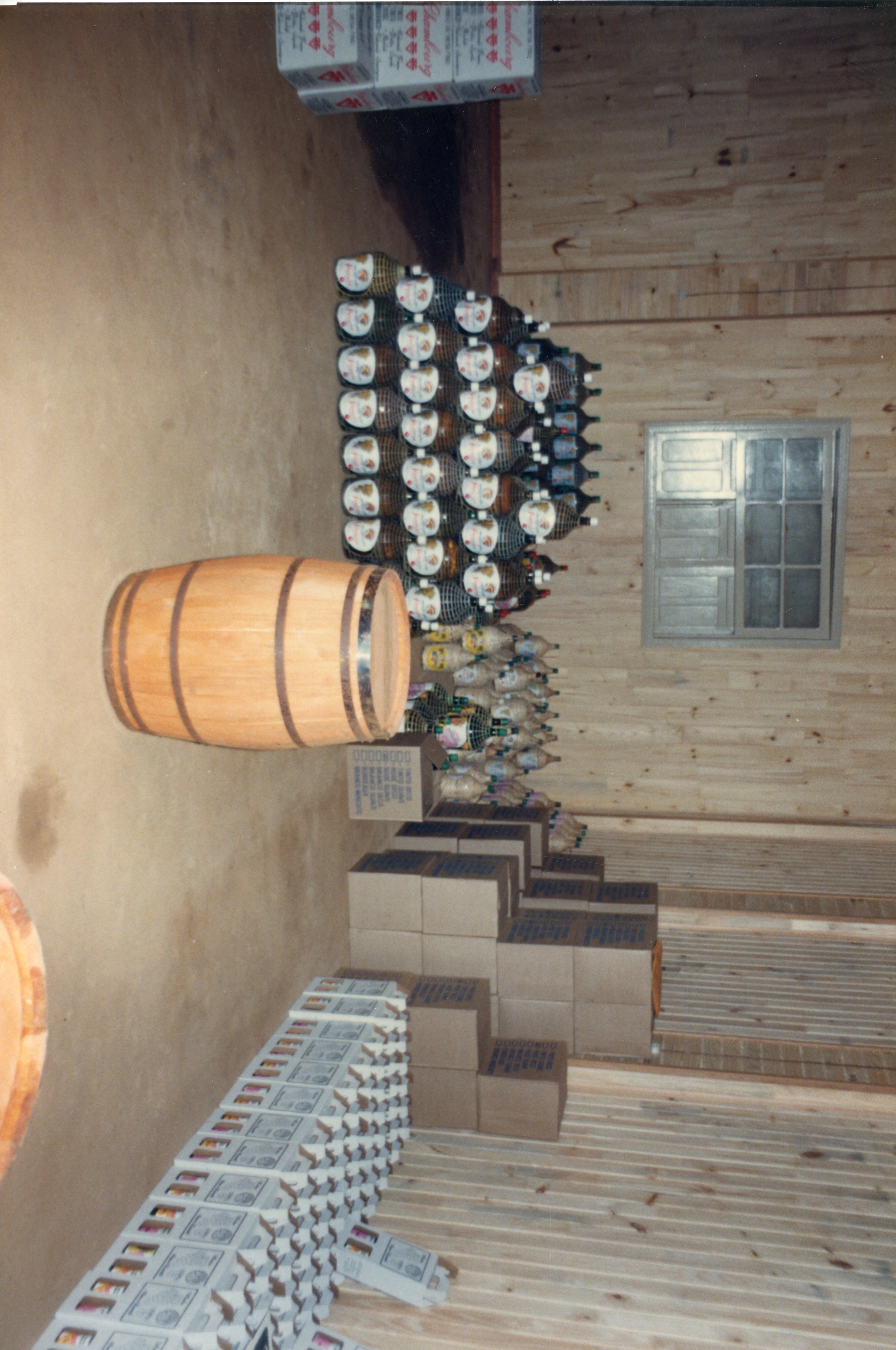 Uma imagem interna mostrando um estoque de bebidas, provavelmente vinho, em um ambiente com paredes de madeira. Observam-se garrafões empilhados, caixas de papelão e um barril de madeira, sugerindo uma vinícola ou adega, comum na região de Videira.
