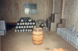 Uma imagem interna mostrando um estoque de bebidas, provavelmente vinho, em um ambiente com paredes de madeira. Observam-se garrafões empilhados, caixas de papelão e um barril de madeira, sugerindo uma vinícola ou adega, comum na região de Videira.