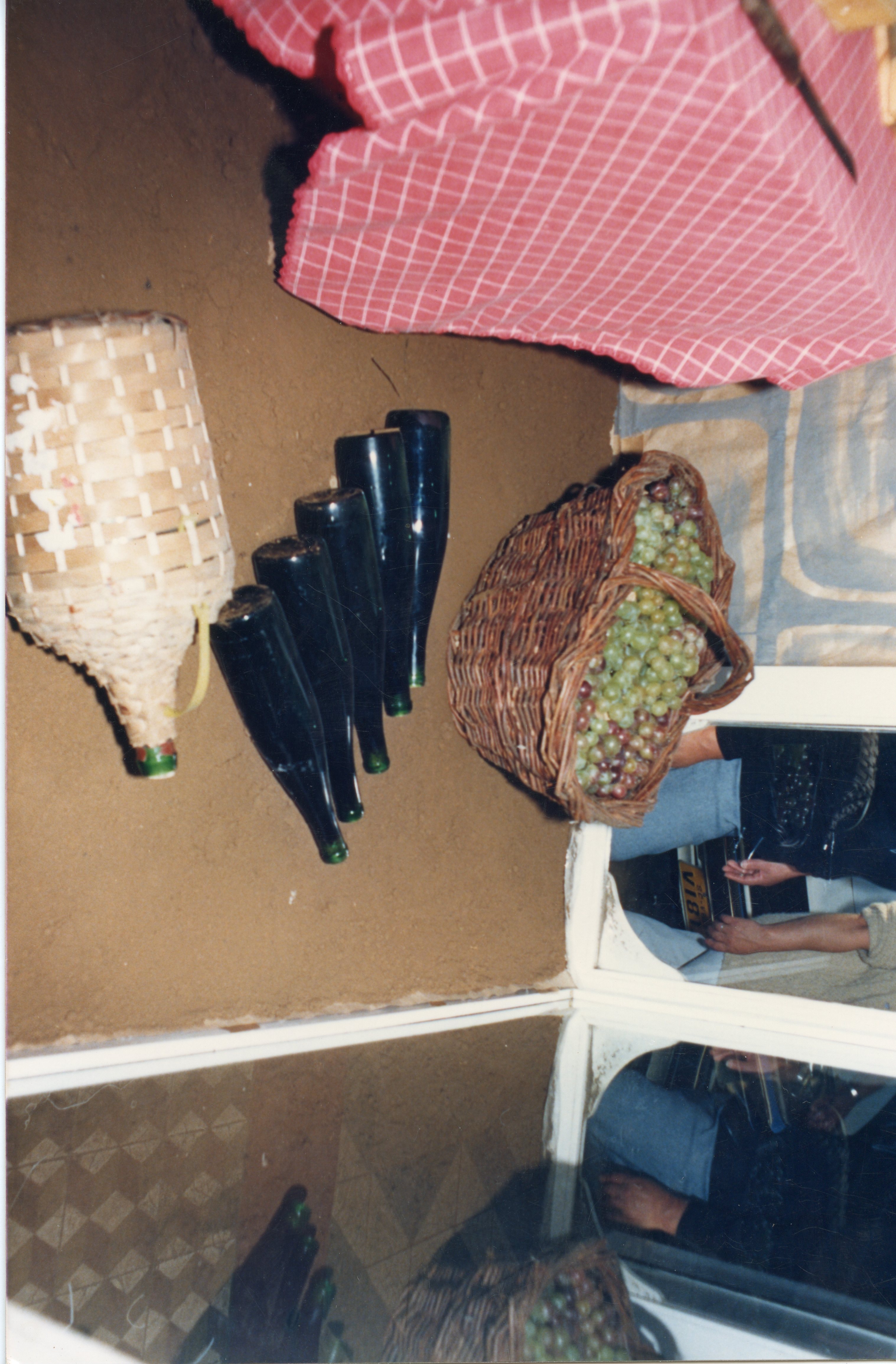 Uma fotografia de ângulo superior mostrando elementos tradicionais da vitivinicultura, incluindo garrafas de vinho escuras, um garrafão revestido de palha e uma cesta de vime cheia de uvas frescas. A cena sugere um contexto rural ou de produção artesanal de vinho, possivelmente em uma região de colonização italiana como Videira, com uma toalha de mesa xadrez vermelha ao fundo.