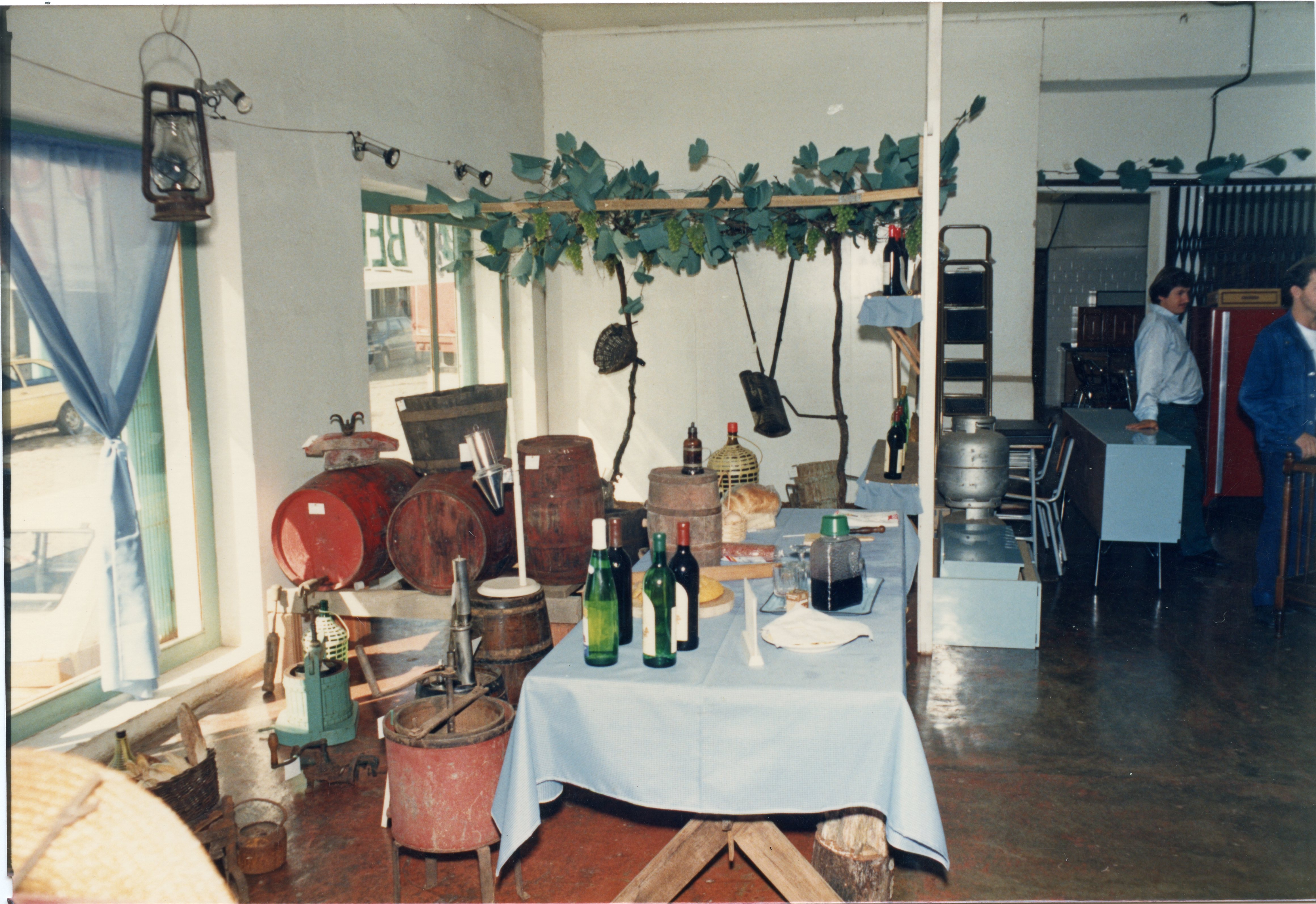 Uma cena interna que parece ser uma exposição ou estande temático sobre a cultura da vitivinicultura, possivelmente em Videira, Santa Catarina. O ambiente apresenta barris de madeira, garrafas de vinho, uma prensa antiga e uma parreira decorativa, sugerindo um evento cultural ou comercial relacionado à produção de vinhos, datado provavelmente da década de 1990.