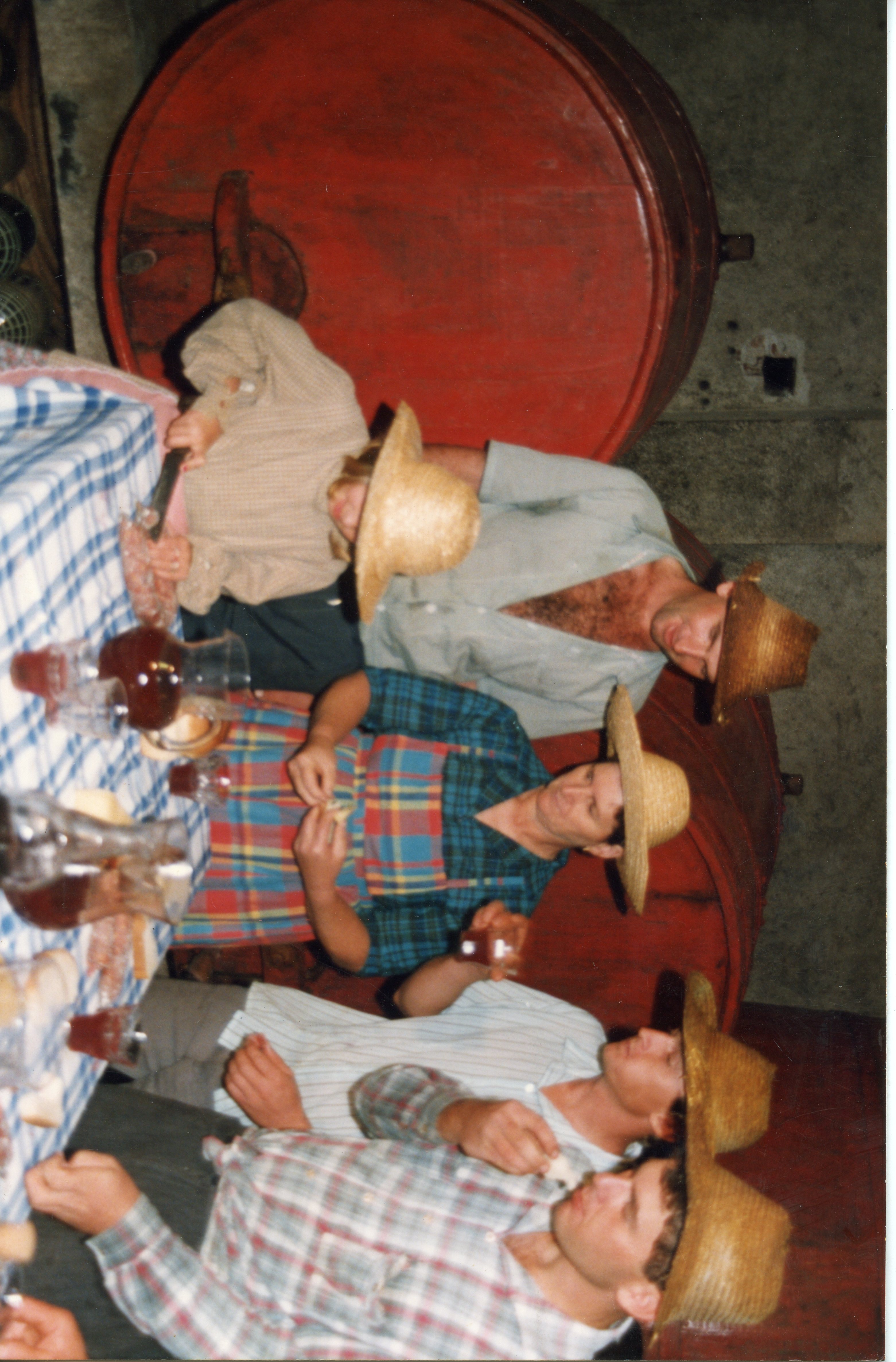 Uma fotografia de um grupo de pessoas em um ambiente interno, possivelmente uma vinícola, com grandes barris de madeira ao fundo. Os indivíduos vestem roupas simples e chapéus de palha, sugerindo uma celebração rural ou colheita de uvas, comum na cultura de imigrantes em regiões como Videira.