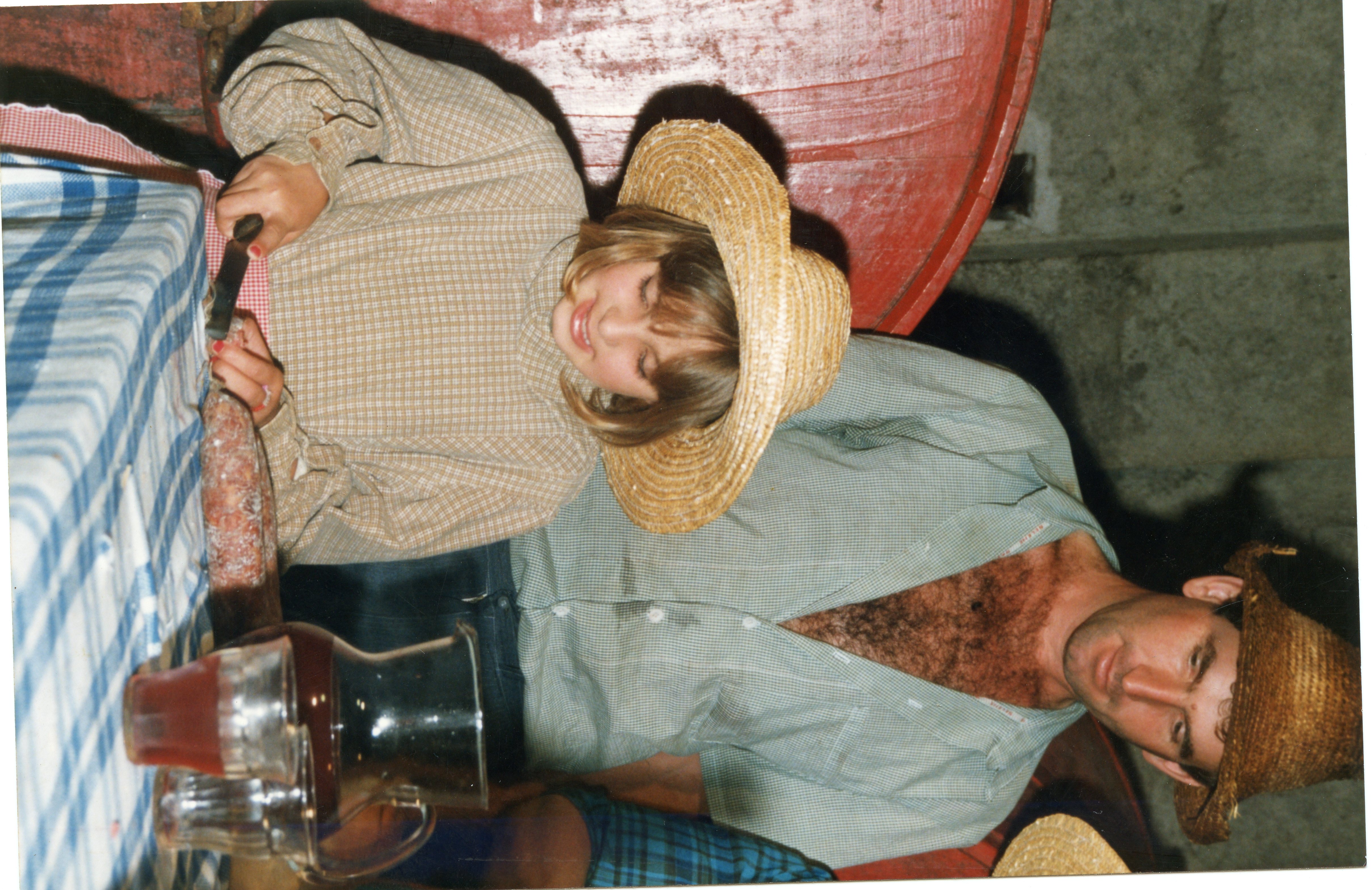 Uma fotografia colorida que mostra uma criança e um homem usando chapéus de palha, sugerindo um contexto rural ou festivo, possivelmente relacionado a festas típicas brasileiras. Eles estão sentados a uma mesa com toalha xadrez, onde há uma jarra de bebida e um alimento, evocando um momento de confraternização familiar.