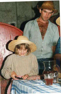 Uma fotografia colorida que mostra uma criança e um homem usando chapéus de palha, sugerindo um contexto rural ou festivo, possivelmente relacionado a festas típicas brasileiras. Eles estão sentados a uma mesa com toalha xadrez, onde há uma jarra de bebida e um alimento, evocando um momento de confraternização familiar.