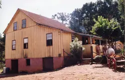 Fotografia de uma casa de madeira tradicional, típica da arquitetura de colonização europeia no sul do Brasil. A estrutura apresenta dois andares, paredes de tábuas verticais e um telhado de duas águas, possivelmente localizada em uma área rural próxima a Videira.