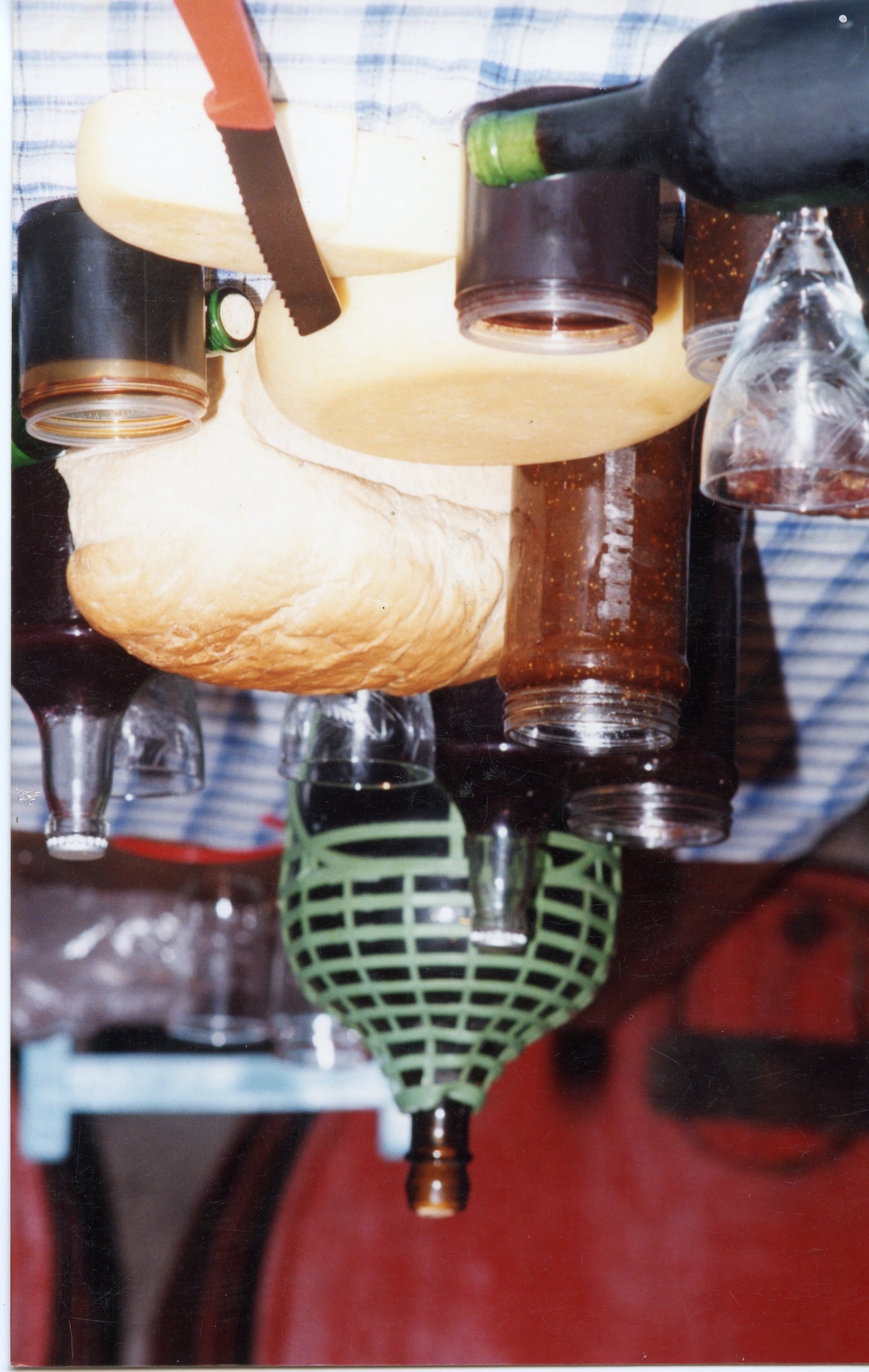Uma fotografia capturada de cima para baixo mostrando uma mesa posta com elementos típicos de uma refeição colonial ou festiva, incluindo pão, queijo, geleias e garrafas de vinho. A presença de uma garrafa com proteção de vime e a disposição dos itens sugerem uma tradição cultural comum em regiões de colonização italiana, como Videira.