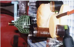 Uma fotografia capturada de cima para baixo mostrando uma mesa posta com elementos típicos de uma refeição colonial ou festiva, incluindo pão, queijo, geleias e garrafas de vinho. A presença de uma garrafa com proteção de vime e a disposição dos itens sugerem uma tradição cultural comum em regiões de colonização italiana, como Videira.