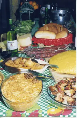 Uma mesa farta preparada para uma refeição festiva, típica da cultura de imigração italiana no sul do Brasil. A cena apresenta pratos tradicionais como polenta, frango, salame, queijos e pães, dispostos sobre uma toalha de mesa xadrez, sugerindo um contexto de celebração familiar ou comunitária.