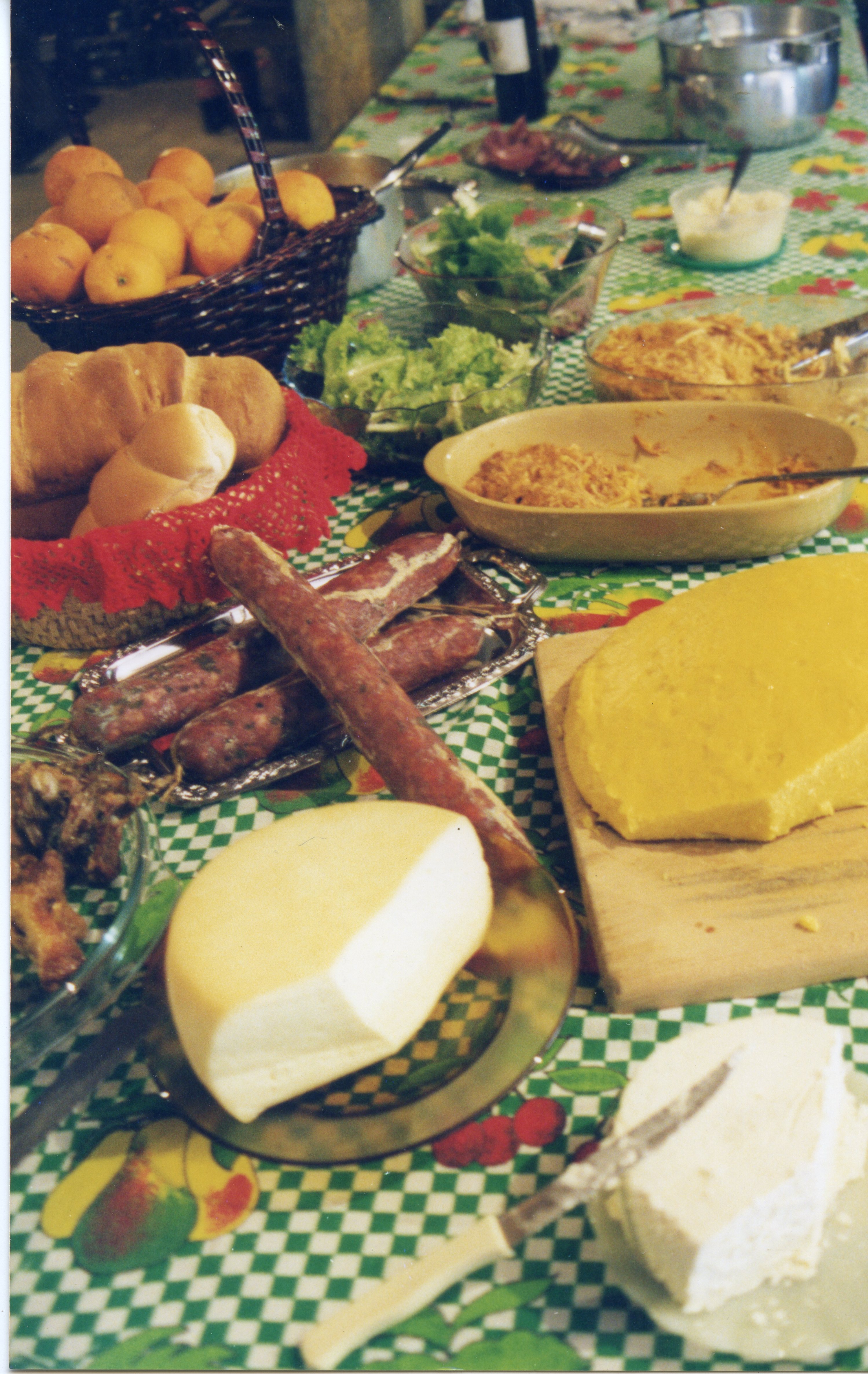 Uma mesa farta preparada para uma refeição, típica de celebrações familiares ou eventos comunitários em regiões de colonização italiana, como Videira. A cena exibe uma variedade de alimentos tradicionais, incluindo queijos, salames, pães, polenta e frutas, dispostos sobre uma toalha de mesa xadrez verde e branca.
