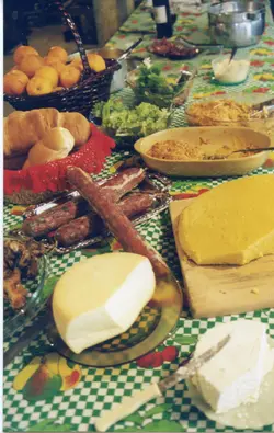 Uma mesa farta preparada para uma refeição, típica de celebrações familiares ou eventos comunitários em regiões de colonização italiana, como Videira. A cena exibe uma variedade de alimentos tradicionais, incluindo queijos, salames, pães, polenta e frutas, dispostos sobre uma toalha de mesa xadrez verde e branca.