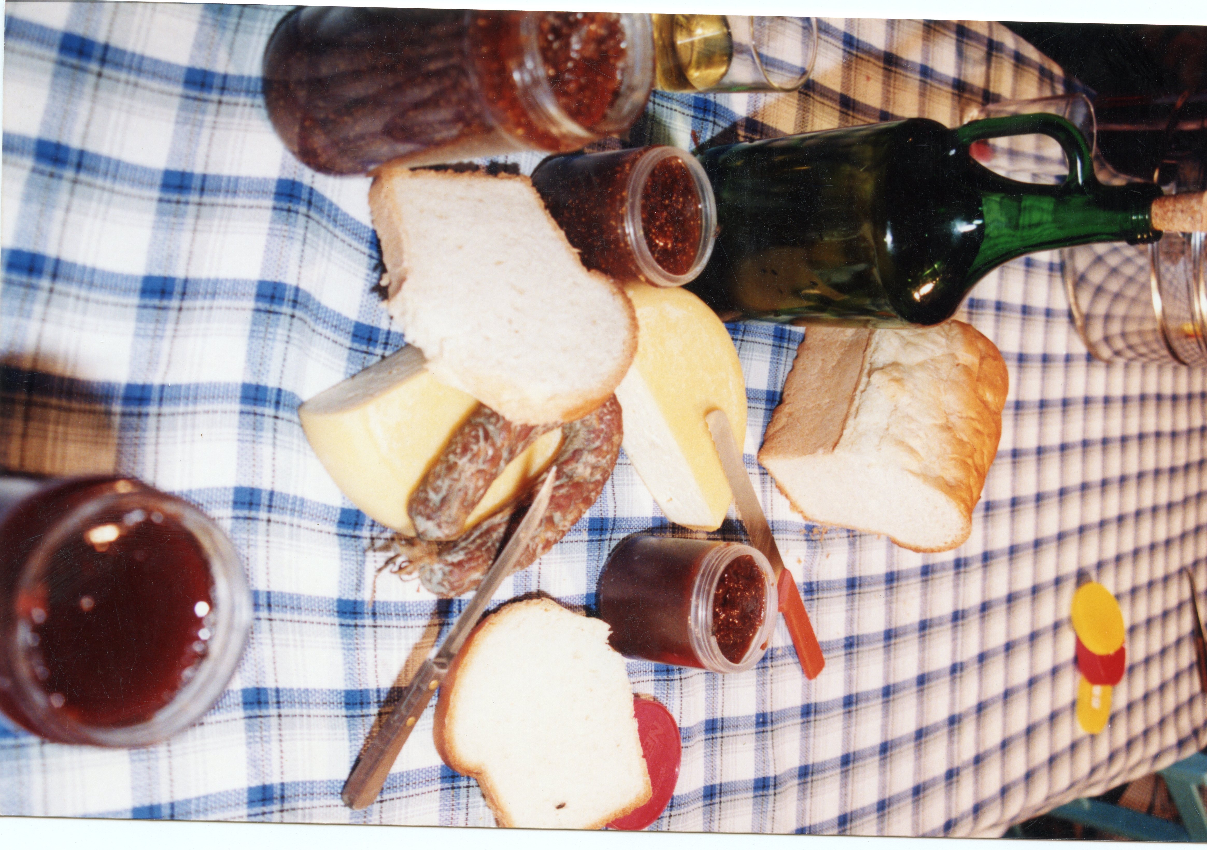 Uma mesa posta com uma toalha xadrez azul e branca, exibindo alimentos típicos de uma refeição colonial ou rural, como pães, queijo, linguiça e compotas em potes de vidro. A cena evoca tradições culinárias comuns em regiões de colonização europeia no Brasil, como Videira, sugerindo um momento de lanche ou café colonial.
