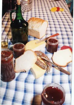 Uma mesa posta com uma toalha xadrez azul e branca, exibindo alimentos típicos de uma refeição colonial ou rural, como pães, queijo, linguiça e compotas em potes de vidro. A cena evoca tradições culinárias comuns em regiões de colonização europeia no Brasil, como Videira, sugerindo um momento de lanche ou café colonial.