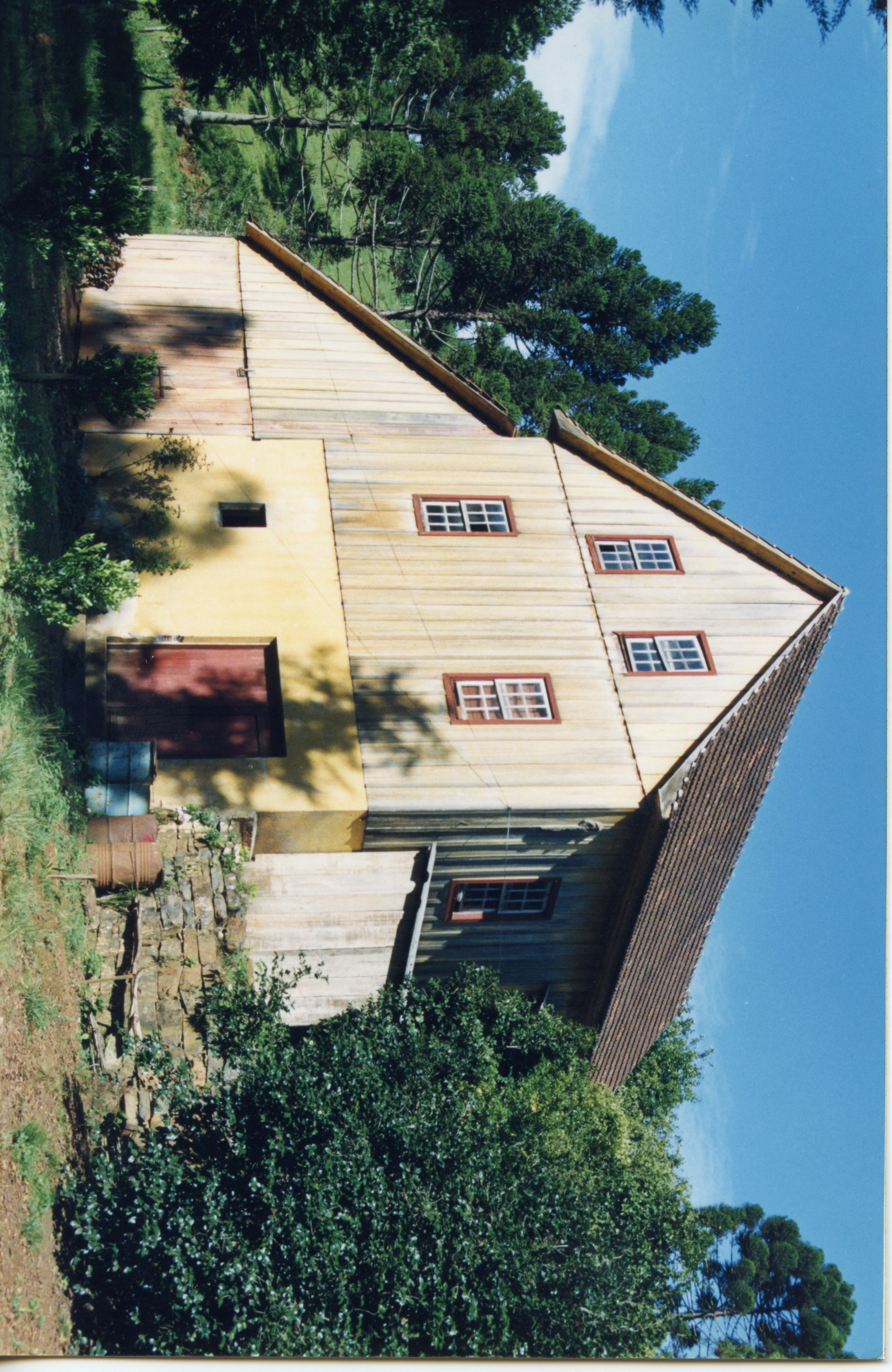 Uma casa de madeira tradicional de dois andares, típica da arquitetura rural de colonização europeia no sul do Brasil. A estrutura apresenta paredes de tábuas verticais, telhado de duas águas e está situada em um ambiente arborizado com araucárias, sugerindo uma paisagem típica da região de Videira.