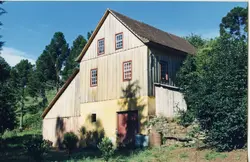 Uma casa de madeira tradicional de dois andares, típica da arquitetura rural de colonização europeia no sul do Brasil. A estrutura apresenta paredes de tábuas verticais, telhado de duas águas e está situada em um ambiente arborizado com araucárias, sugerindo uma paisagem típica da região de Videira.