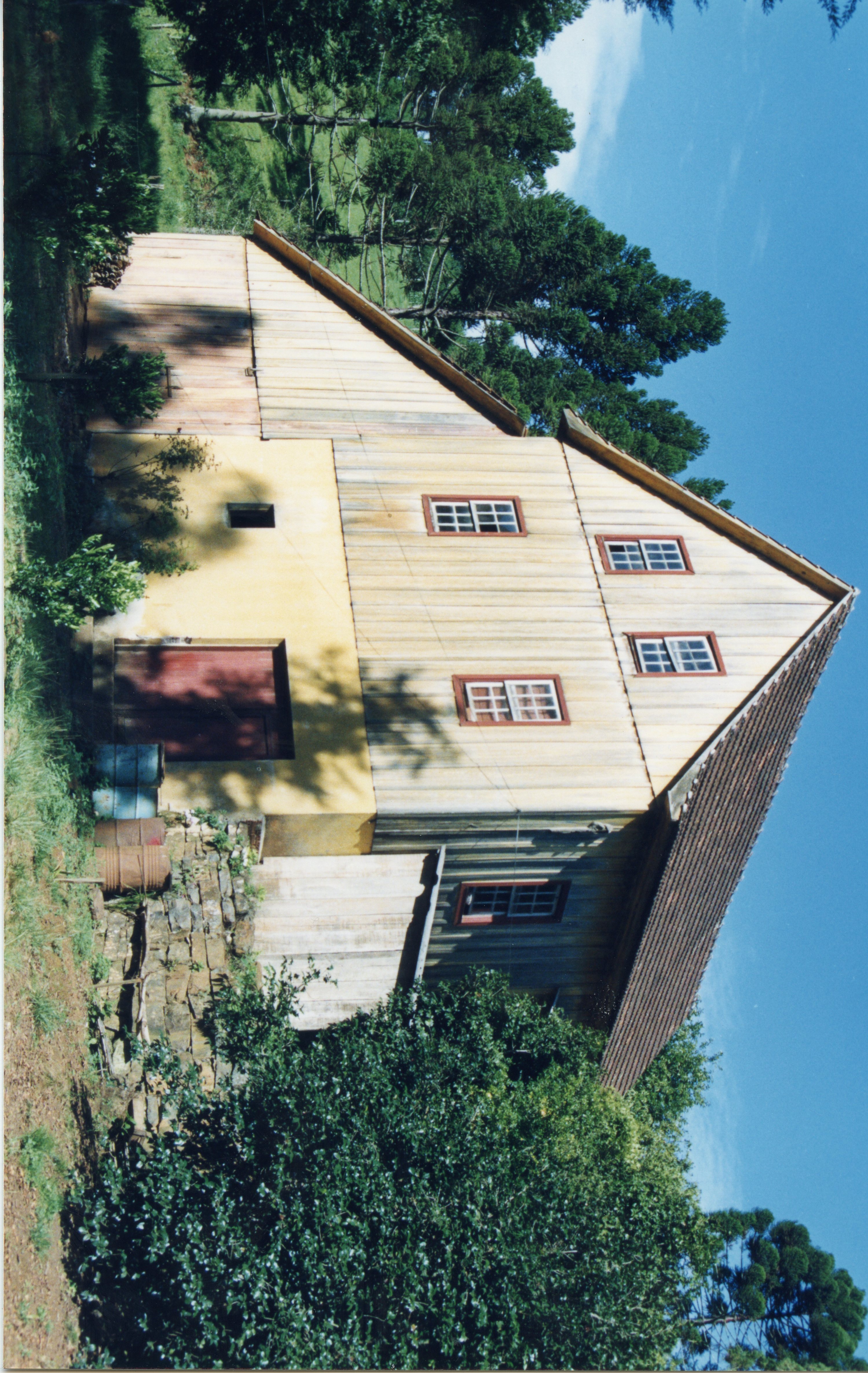 Uma fotografia de uma casa de madeira tradicional, típica da arquitetura de colonização europeia no sul do Brasil. A estrutura apresenta paredes de tábuas verticais, telhado de duas águas e uma base de alvenaria amarela, cercada por vegetação e araucárias, sugerindo um ambiente rural.