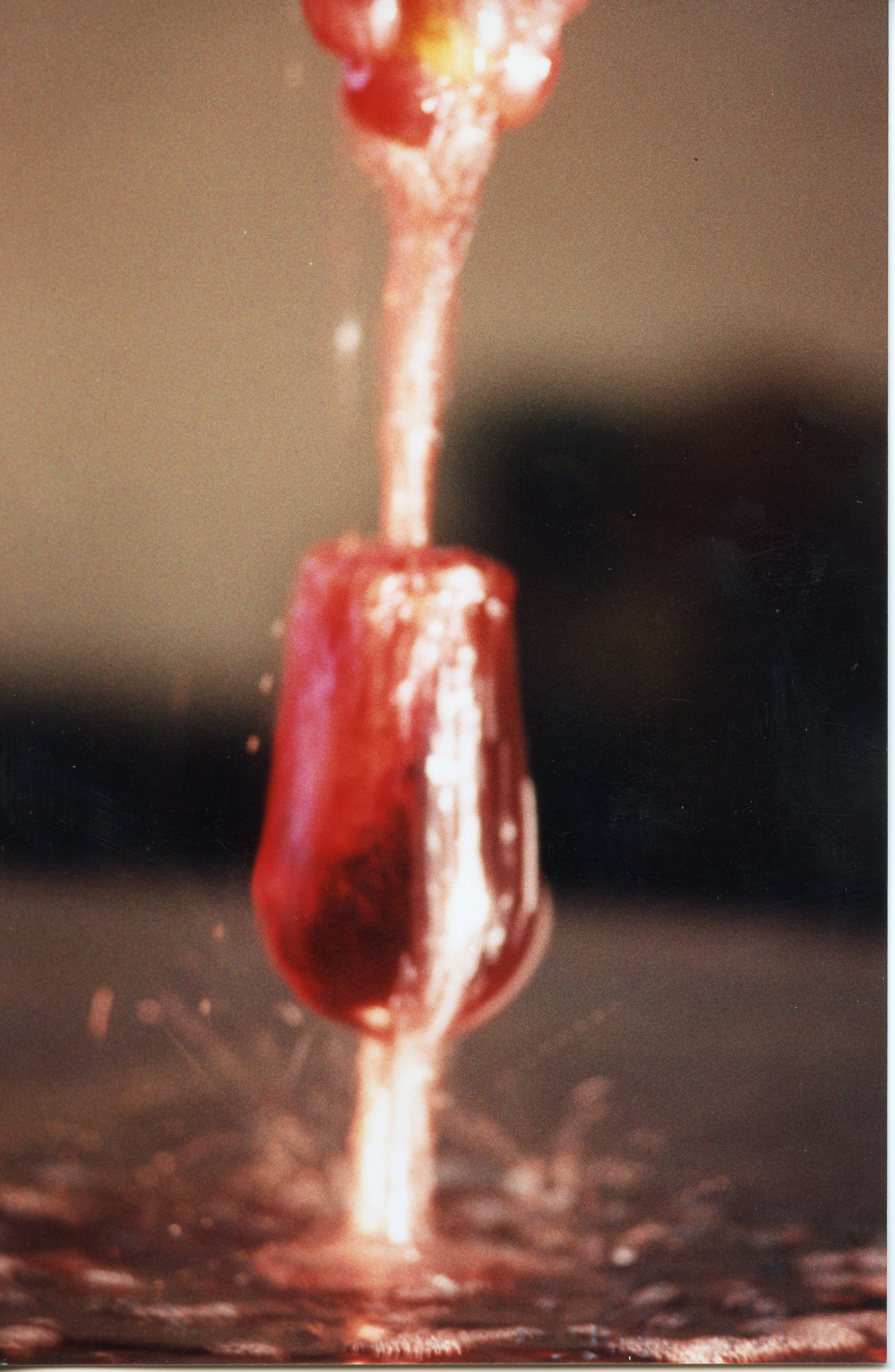 Uma fotografia em close-up capturando o momento dinâmico de um líquido vermelho, possivelmente vinho, sendo derramado em uma taça. A imagem foca no movimento e no respingo do líquido, sugerindo uma técnica de fotografia de alta velocidade, comum em contextos publicitários ou artísticos.