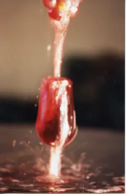 Uma fotografia em close-up capturando o momento dinâmico de um líquido vermelho, possivelmente vinho, sendo derramado em uma taça. A imagem foca no movimento e no respingo do líquido, sugerindo uma técnica de fotografia de alta velocidade, comum em contextos publicitários ou artísticos.