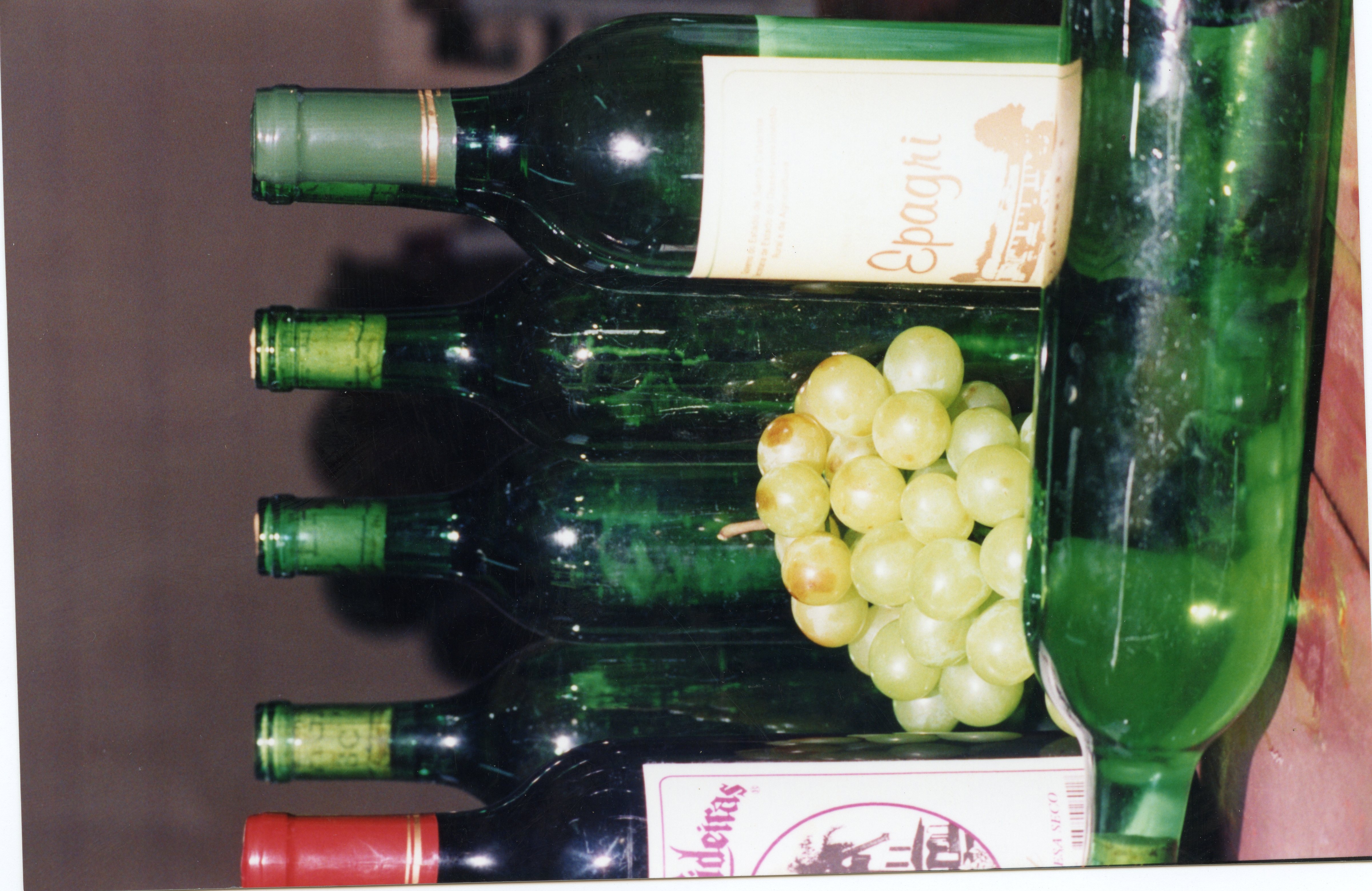 A imagem apresenta várias garrafas de vinho de vidro verde alinhadas horizontalmente, acompanhadas por um cacho de uvas brancas frescas. A presença de rótulos como 'Epagri' e 'Videiras' sugere uma conexão com a vitivinicultura regional, possivelmente em um contexto de promoção de produtos locais.