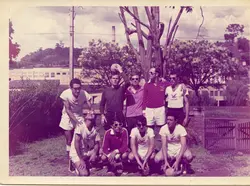 Uma fotografia de grupo de um time de futebol amador posando ao ar livre, provavelmente na década de 1970 ou 1980. Os jogadores vestem uniformes esportivos, alguns usam óculos escuros, e o cenário inclui uma área gramada com uma construção industrial ao fundo, sugerindo um contexto de lazer em uma cidade do interior.