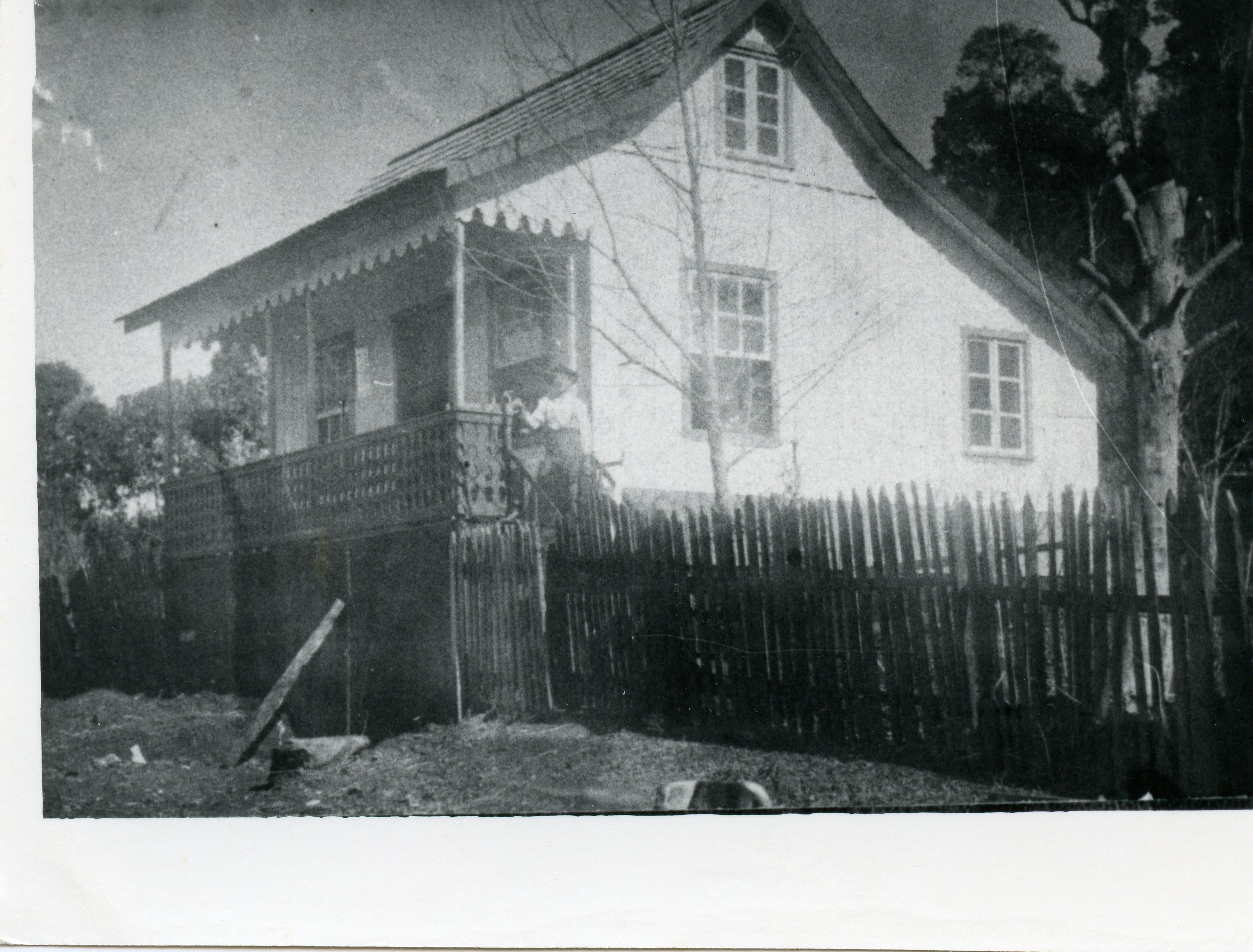 Fotografia antiga em preto e branco retratando uma residência de madeira com varanda decorada e cerca de estacas de madeira. A imagem sugere um contexto de colonização rural ou urbana inicial, possivelmente na primeira metade do século XX, com uma pessoa visível na varanda.