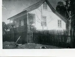 Fotografia antiga em preto e branco retratando uma residência de madeira com varanda decorada e cerca de estacas de madeira. A imagem sugere um contexto de colonização rural ou urbana inicial, possivelmente na primeira metade do século XX, com uma pessoa visível na varanda.