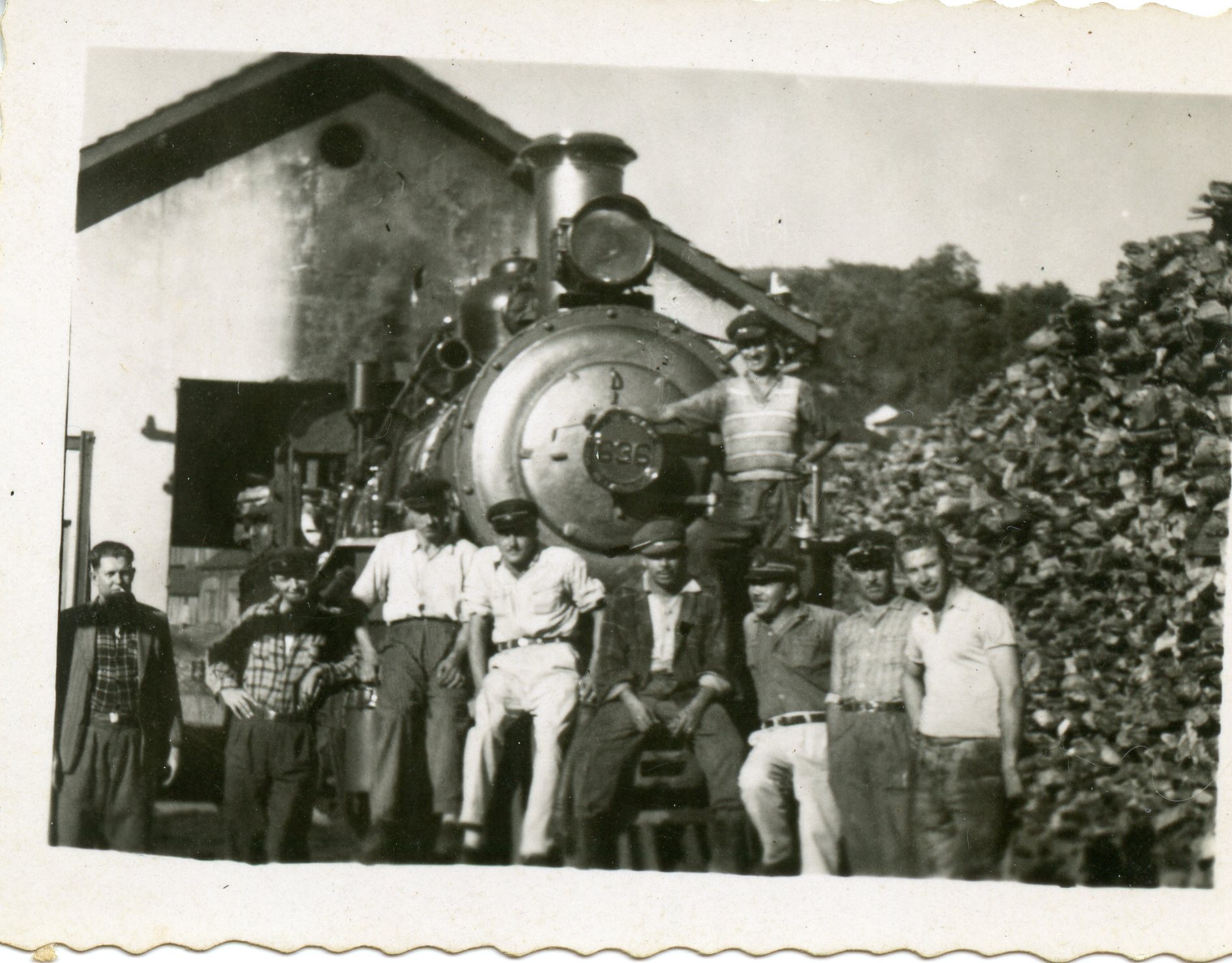 Fotografia em preto e branco retratando um grupo de trabalhadores ferroviários posando em frente a uma locomotiva a vapor de grande porte. A cena sugere um ambiente de manutenção ou pátio de manobras, com uma pilha de lenha ou carvão ao lado, típica da infraestrutura ferroviária do início ao meados do século XX no Brasil.