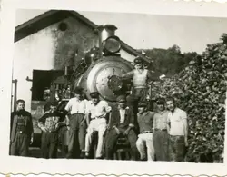Fotografia em preto e branco retratando um grupo de trabalhadores ferroviários posando em frente a uma locomotiva a vapor de grande porte. A cena sugere um ambiente de manutenção ou pátio de manobras, com uma pilha de lenha ou carvão ao lado, típica da infraestrutura ferroviária do início ao meados do século XX no Brasil.