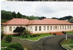Uma fotografia colorida de um edifício térreo de arquitetura institucional, possivelmente uma escola ou prédio público, com telhado de telhas cerâmicas e paredes em tons pastéis. O cenário apresenta um jardim bem cuidado e um caminho pavimentado, sugerindo uma construção de meados do século XX em uma região de colonização europeia no Brasil.