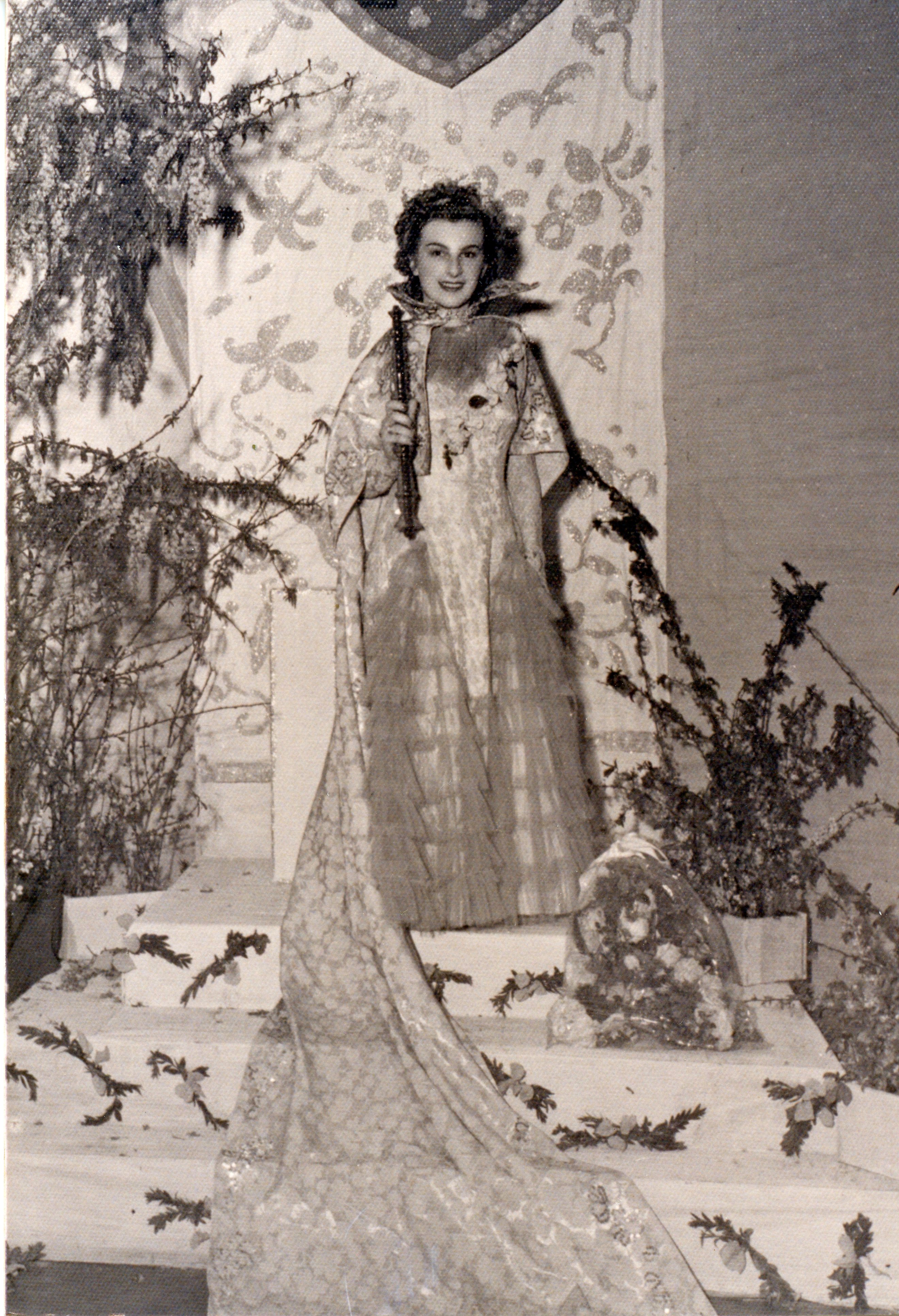 Uma fotografia em preto e branco retratando uma jovem vestida com trajes de gala, possivelmente uma rainha de festividades ou concurso de beleza local. Ela está em um palco decorado com elementos florais e um fundo ornamentado, segurando um cetro, sugerindo um evento social ou comemorativo de meados do século XX.