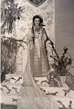 Uma fotografia em preto e branco retratando uma jovem vestida com trajes de gala, possivelmente uma rainha de festividades ou concurso de beleza local. Ela está em um palco decorado com elementos florais e um fundo ornamentado, segurando um cetro, sugerindo um evento social ou comemorativo de meados do século XX.