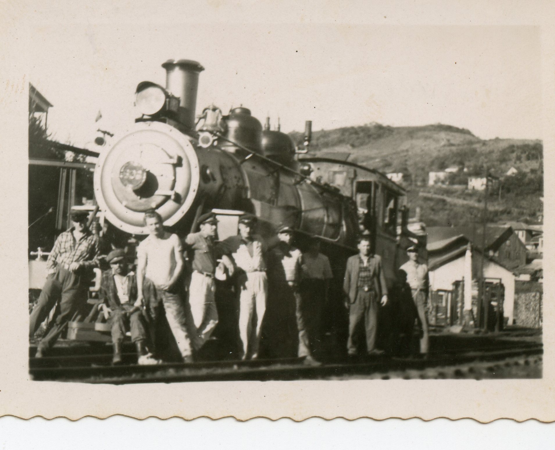 Fotografia em preto e branco retratando um grupo de homens posando em frente a uma locomotiva a vapor de grande porte. O cenário sugere uma área ferroviária em uma pequena cidade brasileira, com casas ao fundo em uma encosta, possivelmente datando da metade do século XX.