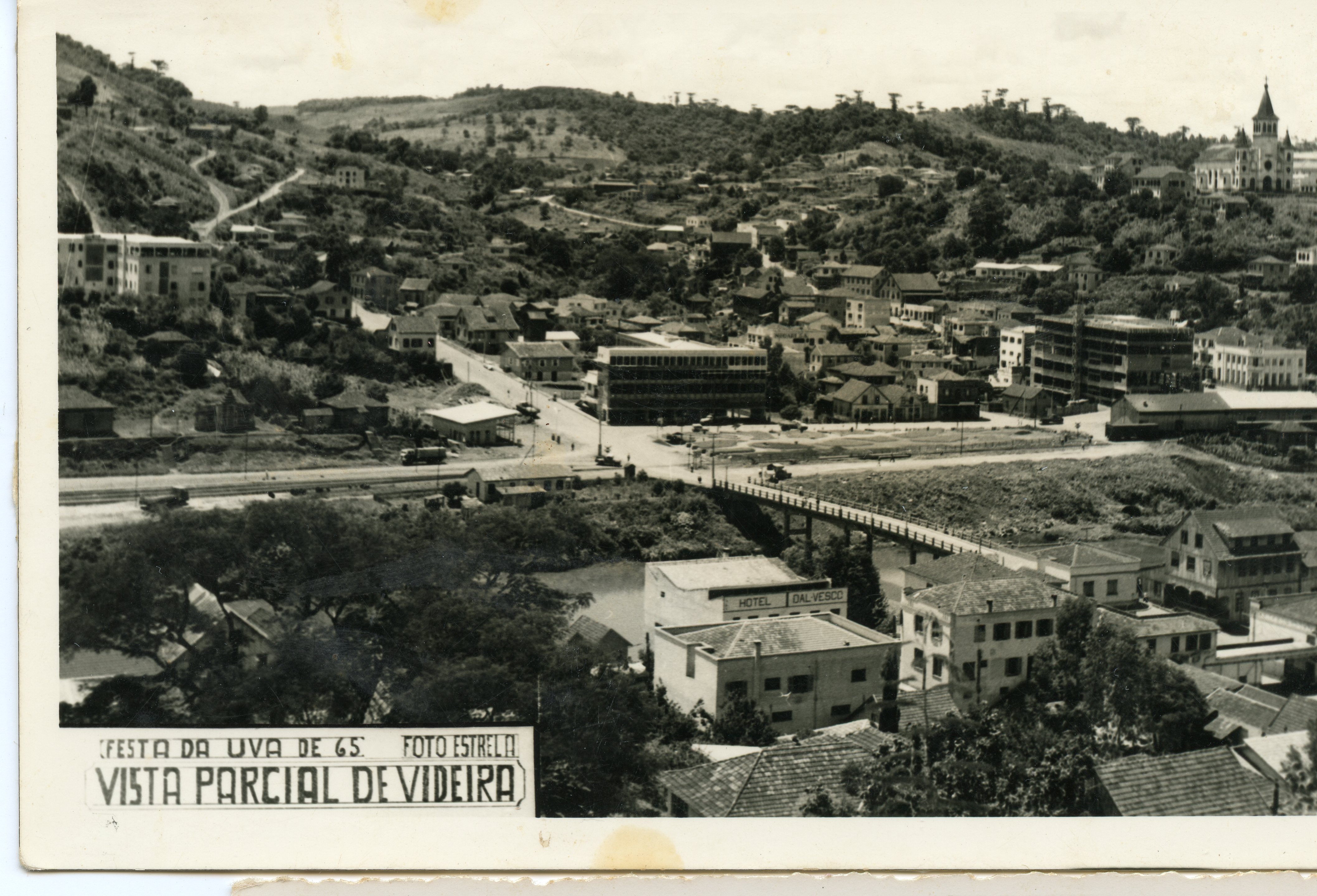 Fotografia panorâmica em preto e branco da cidade de Videira, Santa Catarina, datada de 1965 durante a Festa da Uva. A imagem exibe o desenvolvimento urbano da época, com destaque para a igreja, a ponte sobre o rio e o Hotel Dal Vesco, capturando a paisagem característica da região.