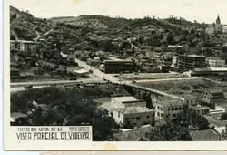 Fotografia panorâmica em preto e branco da cidade de Videira, Santa Catarina, datada de 1965 durante a Festa da Uva. A imagem exibe o desenvolvimento urbano da época, com destaque para a igreja, a ponte sobre o rio e o Hotel Dal Vesco, capturando a paisagem característica da região.