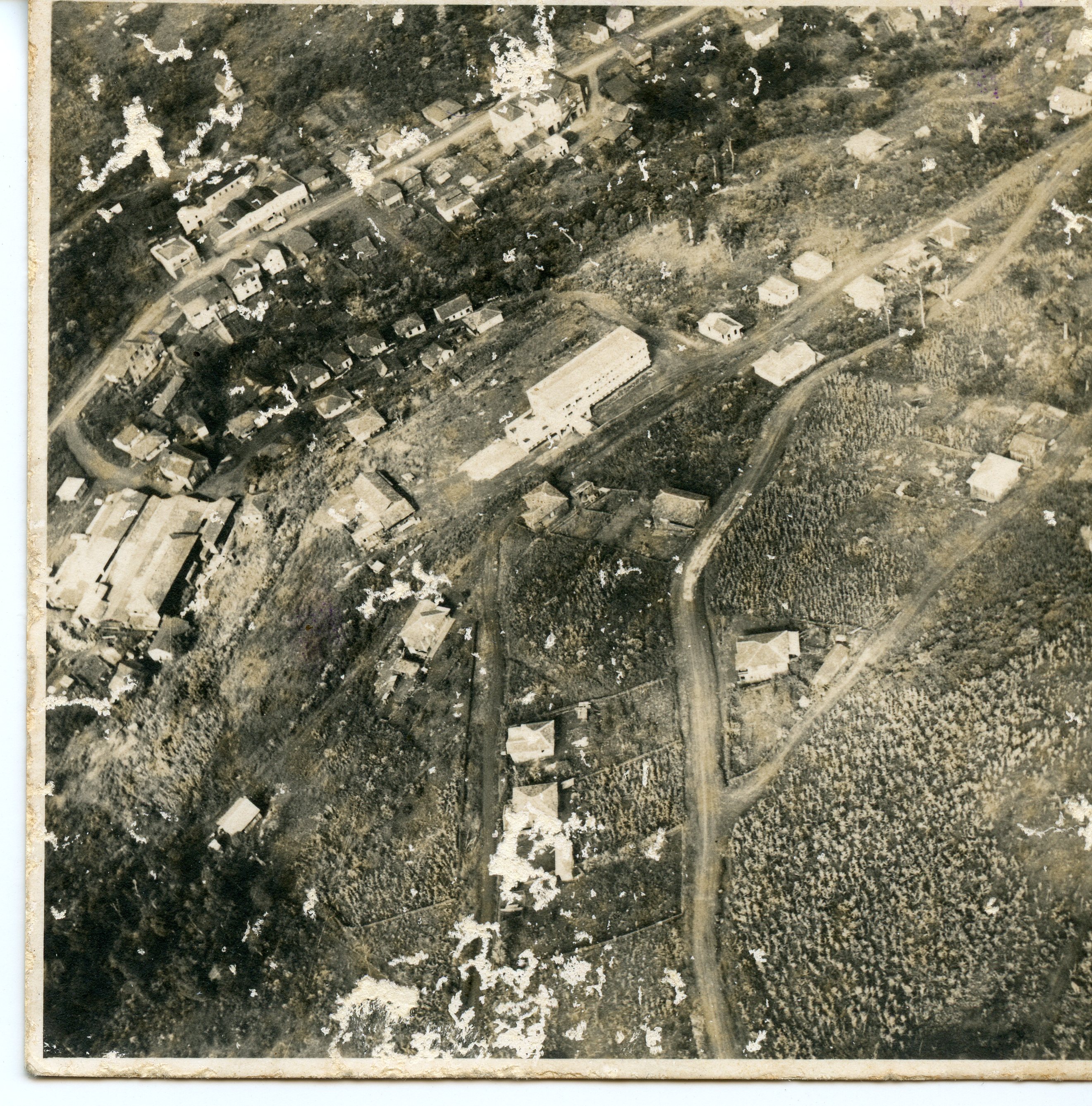 Fotografia aérea em preto e branco mostrando o desenvolvimento urbano inicial de uma pequena localidade, possivelmente no interior do Brasil. A imagem exibe casas dispersas, estradas de terra e grandes edificações que sugerem uma estrutura industrial ou institucional, refletindo o período de expansão e colonização da região.